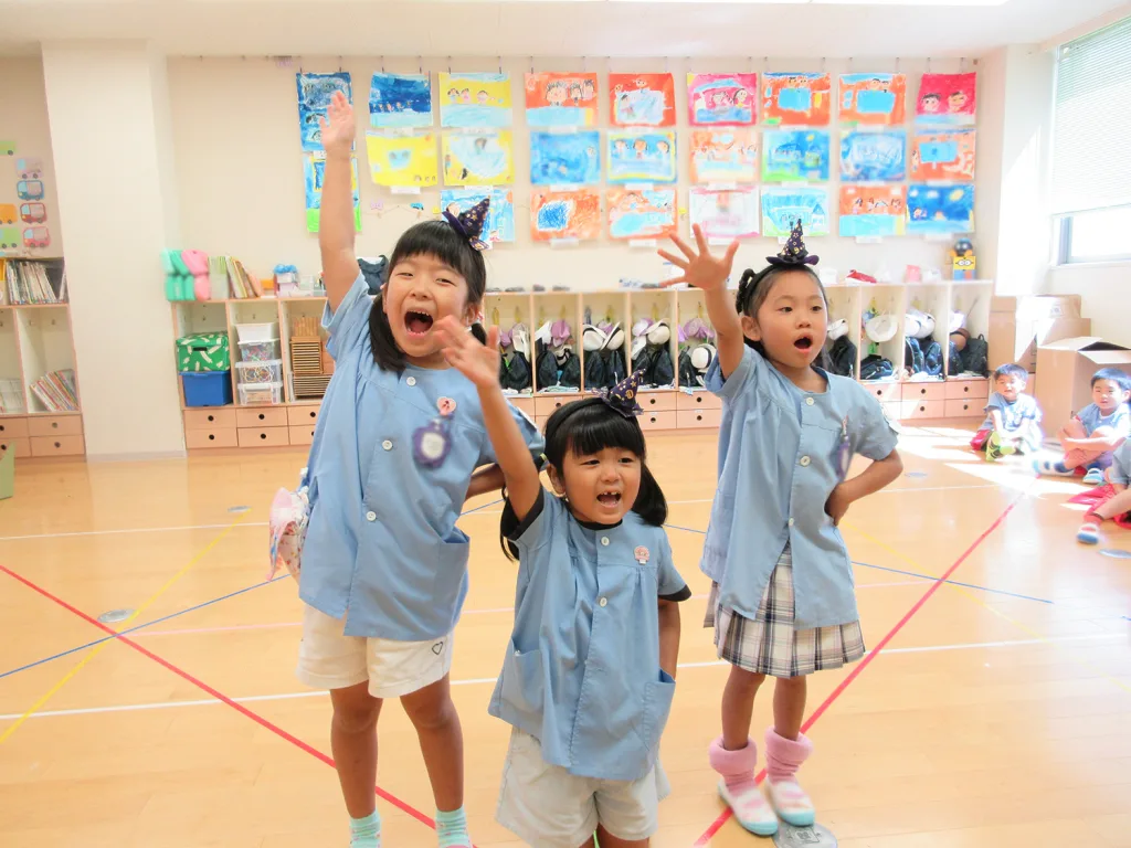 Three smiling children in blue smocks cheer with arms raised during a classroom activity, colorful artwork on the wall behind them.