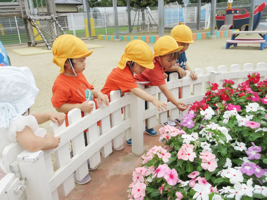 Young children in orange shirts and yellow hats lean over a white fence to look at pink and white flowers in a garden bed.
