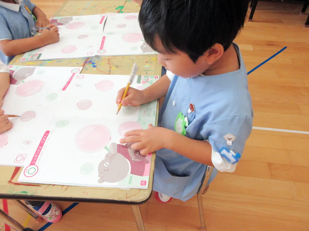 Young child in a light blue smock drawing with a yellow pencil at a classroom table covered with worksheets featuring large pink circles.