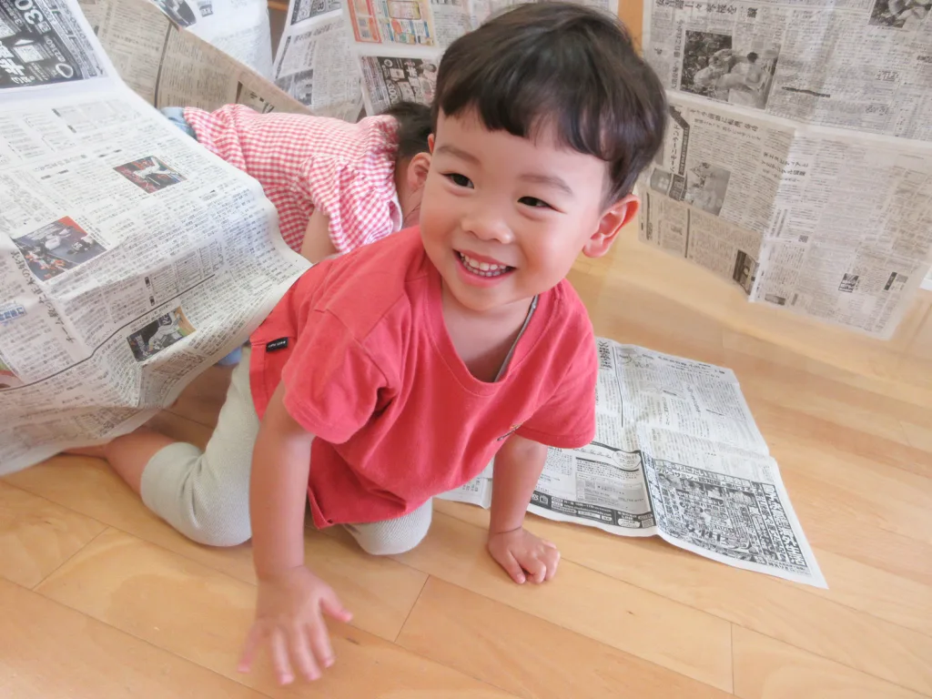 Smiling toddler in a red shirt crawling on a wooden floor, surrounded by hanging newspaper sheets like a makeshift tunnel.