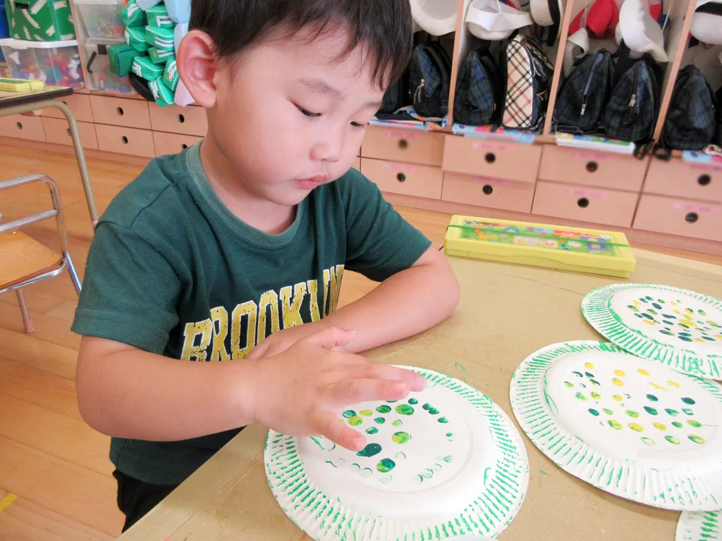 Close-up of a young boy stamping green dots onto a paper plate during art class in a classroom.
