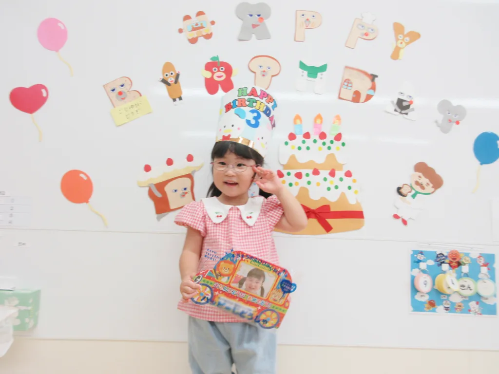 Young girl in a pink gingham shirt wears a tall birthday hat that says 'Happy Birthday 3' and poses with a colorful prize, in front of a wall decorated with balloons and cake decorations.