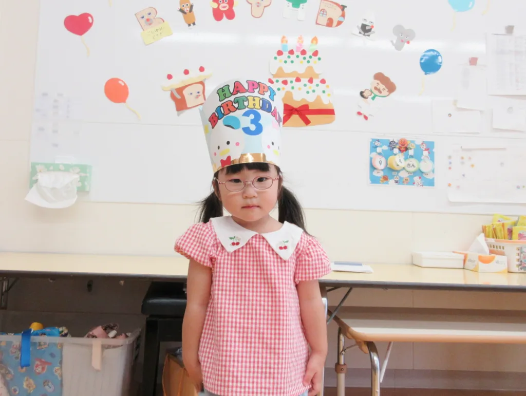 Young girl in a pink gingham dress wears a tall paper birthday crown that reads 'Happy Birthday 3' in a decorated classroom backdrop.