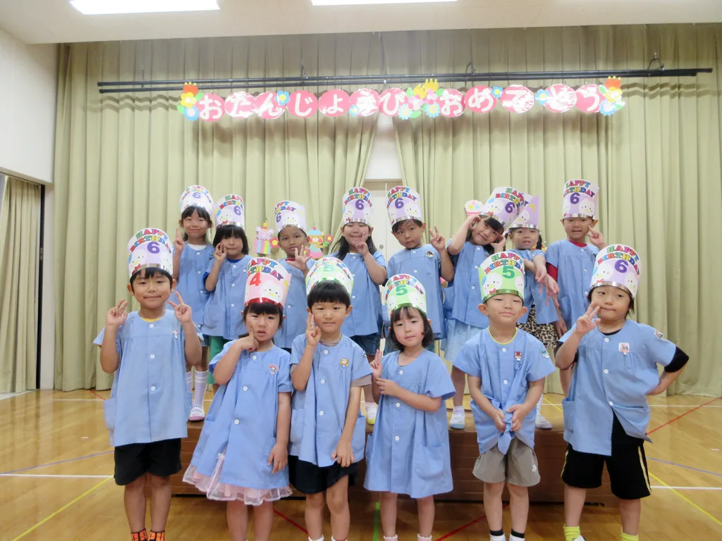 Group of young children in blue uniforms posing on a stage, wearing colorful birthday hats and making peace signs during a celebration. A banner with Japanese text hangs above them.