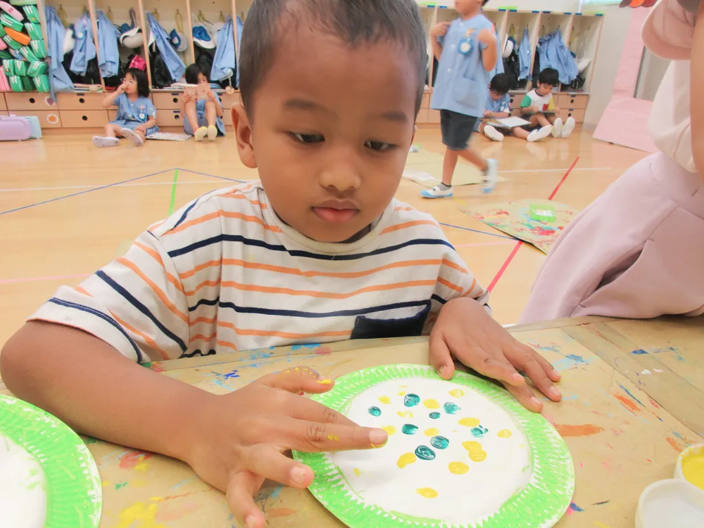 Child at a classroom art table pressing yellow and blue dots onto a paper plate.",