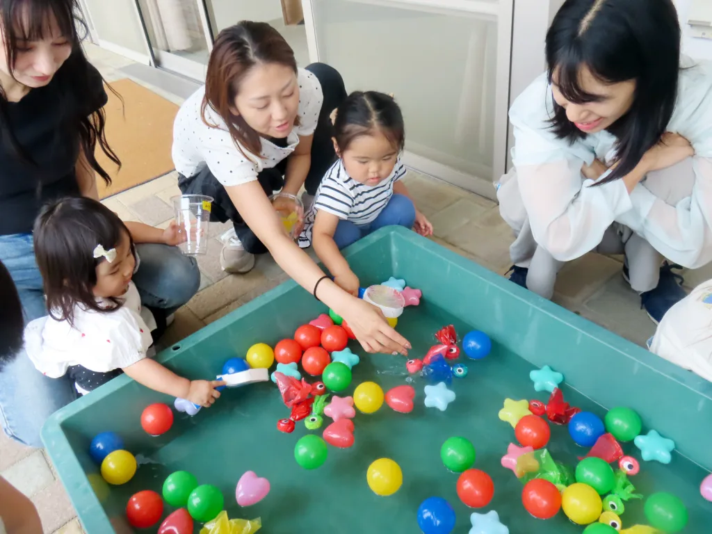 Several children and adults crouch beside a teal water table filled with colorful floating toys and balls, engaging in a show-and-tell water play.