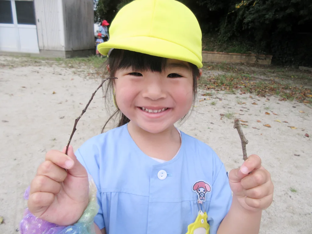 Smiling young girl in a bright yellow cap holding two small sticks outdoors on a sandy ground.