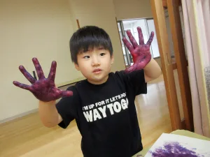 Young child with purple paint on both hands, wearing a black shirt, showing hands to the camera in a classroom setting. The shirt reads: 'WAY TO GO' (partial visible).