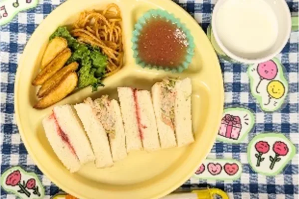 Yellow divided plate with finger sandwiches, potato wedges, greens, noodles, a jelly cup, and a cup of milk on a blue check tablecloth.