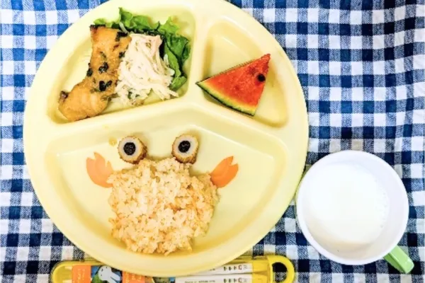 Divided yellow plate with crab-shaped rice, olive eyes, carrot claws, fried chicken with greens, a watermelon slice, and a cup of milk for a child lunch.
