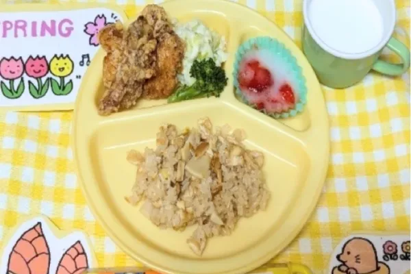 Three-compartment yellow plate with fried chicken and greens, shredded chicken on the bottom, a strawberry dessert cup, and a mug of milk on a yellow checkered tablecloth.