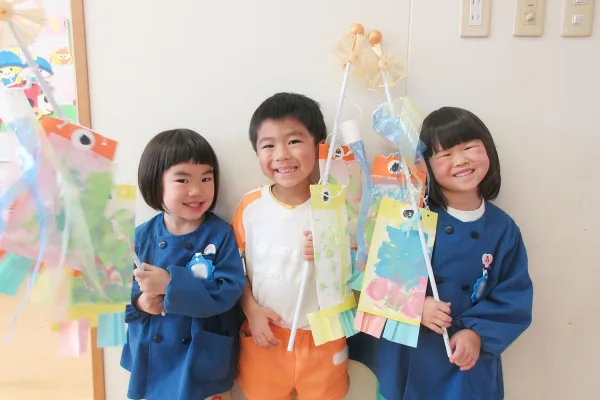 Three smiling children pose together, holding colorful paper crafts on sticks and wearing bright blue coats (center child in orange shorts).