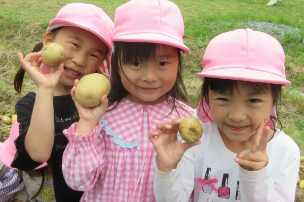 Three young girls wearing pink hats hold potatoes they harvested in a green field.