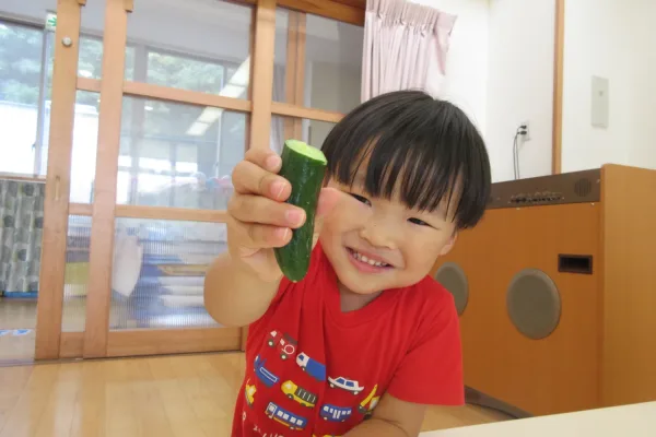 Smiling young child in a red shirt holds up a green cucumber toward the camera indoors, showing the cut end of the cucumber.