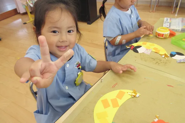 A smiling preschool girl in a blue smock holds up a peace sign toward the camera while crafting at a table with yellow paper mosaics.