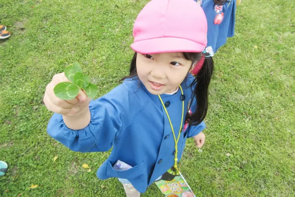 Young girl in a pink cap offering a four-leaf clover outdoors on grass field, wearing a blue jacket and a yellow lanyard.