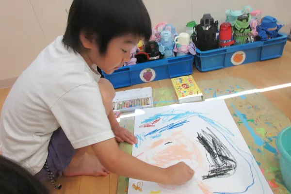 Young child kneels on the floor, drawing with crayons on a large sheet of paper with colorful scribbles.