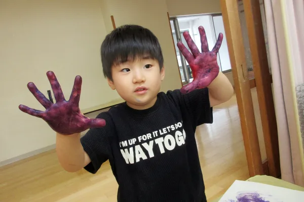 Young child with purple paint on both hands, wearing a black shirt, showing hands to the camera in a classroom setting. The shirt reads: 'WAY TO GO' (partial visible).